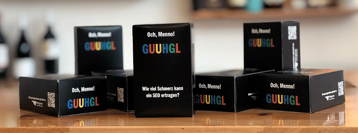 Mehrere schwarze Boxen mit der Aufschrift „Och, Menno! GUUHGL – Wie viel Schmerz kann ein SEO ertragen?“ stehen auf einem Holztisch. Im Hintergrund sind unscharf Flaschen und Gläser in einem Regal zu sehen, die eine gemütliche Bar- oder Büroumgebung andeuten.