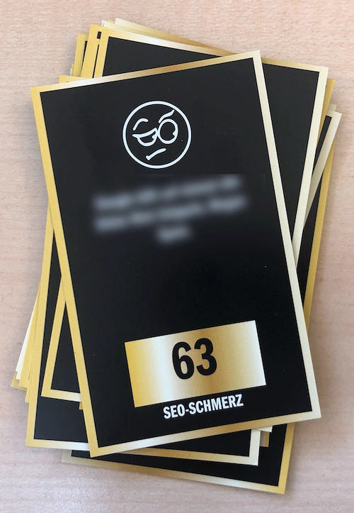 Schwarze Karte mit goldfarbenem Rand, oben ein stilisiertes Gesicht mit Monokel, unten die Zahl '63' und der Text 'SEO-SCHMERZ'