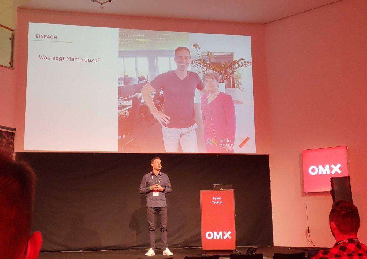 Franz Tretter auf der OMX-Bühne, auf der Leinwand hinter ihm die Folie „EINFACH – Was sagt Mama dazu?“ mit einem Foto von ihm und seiner Papp-Mama