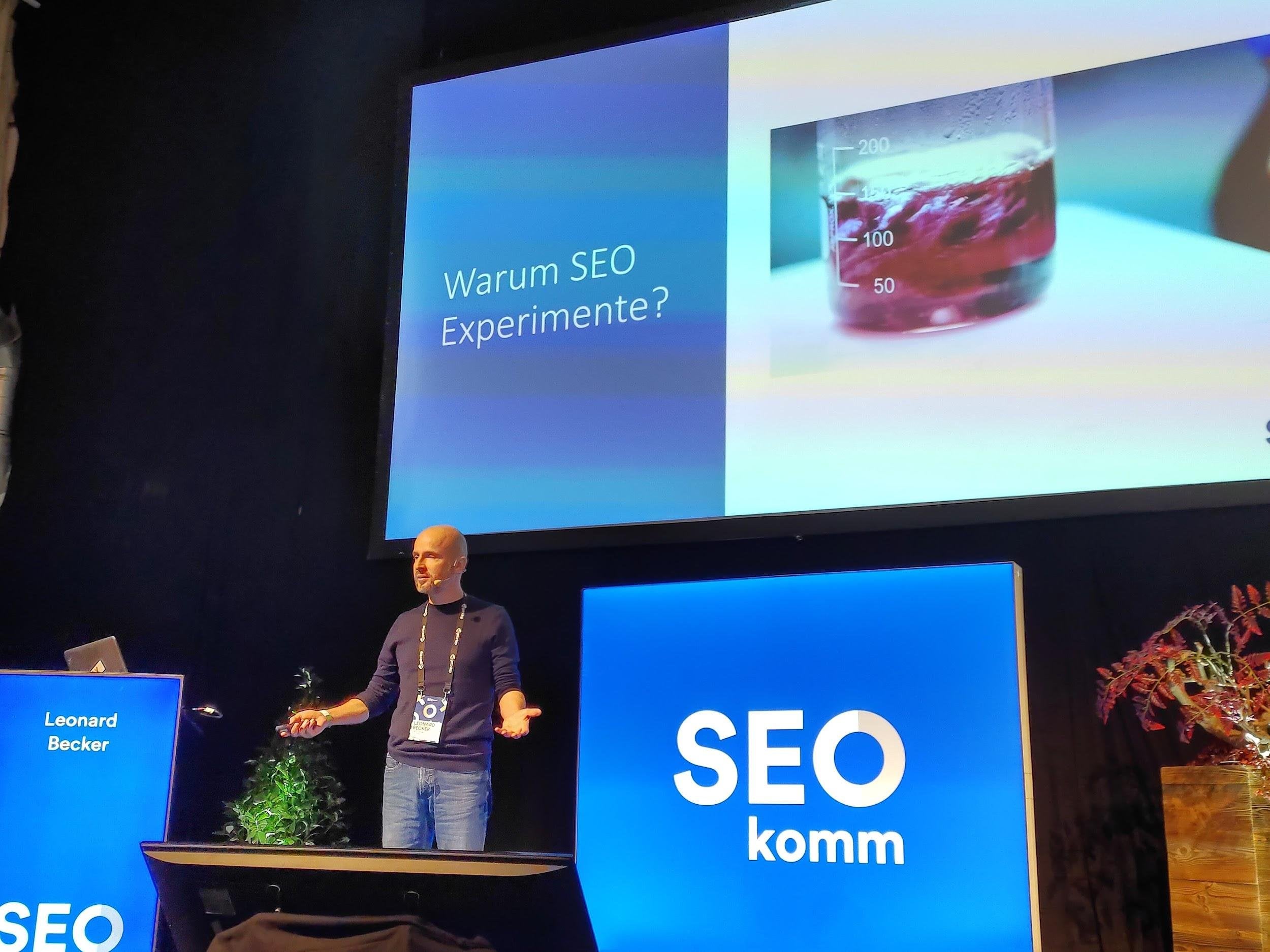 Leonard Becker steht auf der SEOkomm-Bühne vor einer Leinwand mit der Folie „Warum SEO Experimente?“, rechts daneben ein Becherglas mit roter Flüssigkeit.