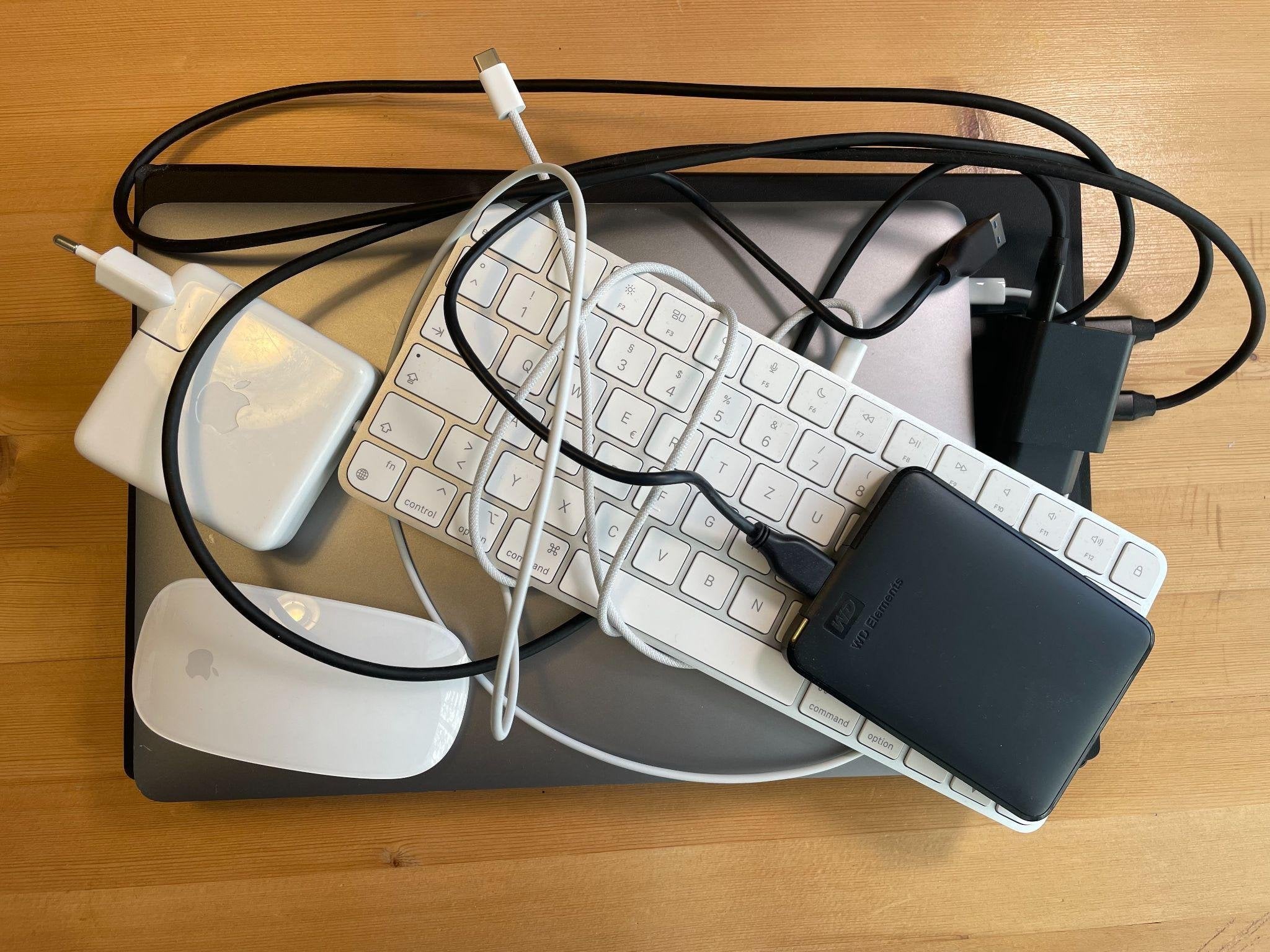 Auf einem geschlossenen Laptop stapeln sich Tastatur, Maus, Netzteil, eine externe Festplatte und aufgerollte Kabel – das zusammengepackte Home-Office-Equipment.