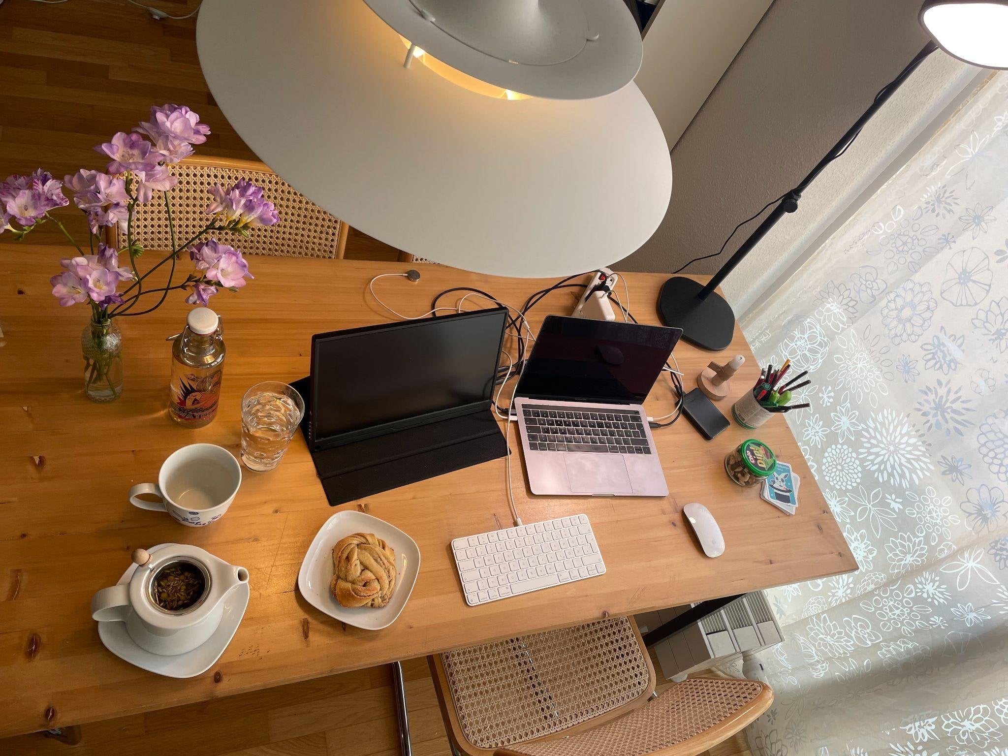 Blick von oben auf einen Holztisch mit mobilem Zusatzmonitor, Laptop, Tastatur und Maus, daneben Teekanne, Tasse, eine Zimtschnecke und ein Strauß Freesien.