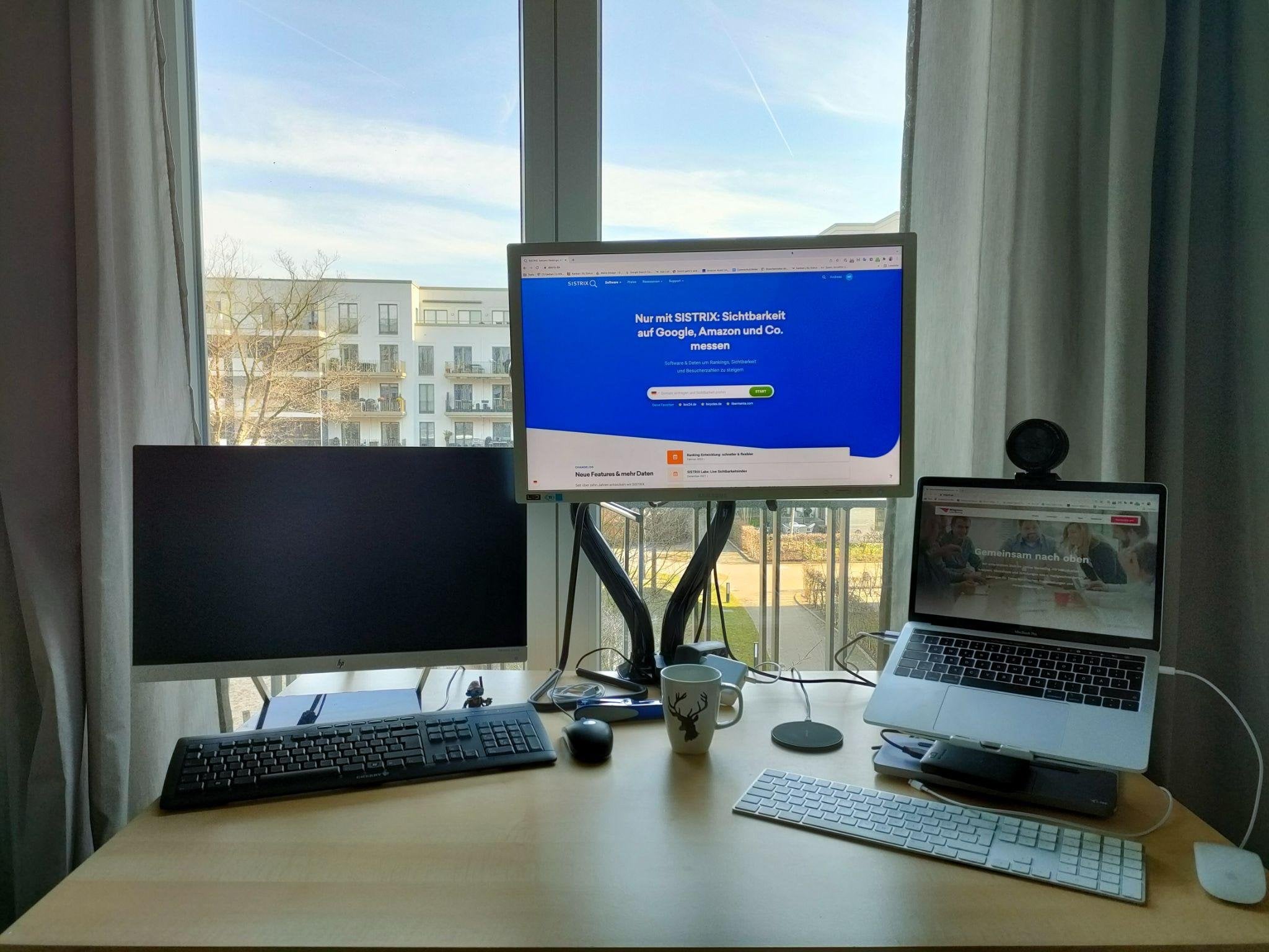 Schreibtisch am Fenster: Ein großer Monitor am Schwenkarm zeigt die Sistrix-Startseite, links steht ein zweiter, dunkler Bildschirm, rechts ein Laptop auf einem Ständer mit aufgesetzter Webcam.