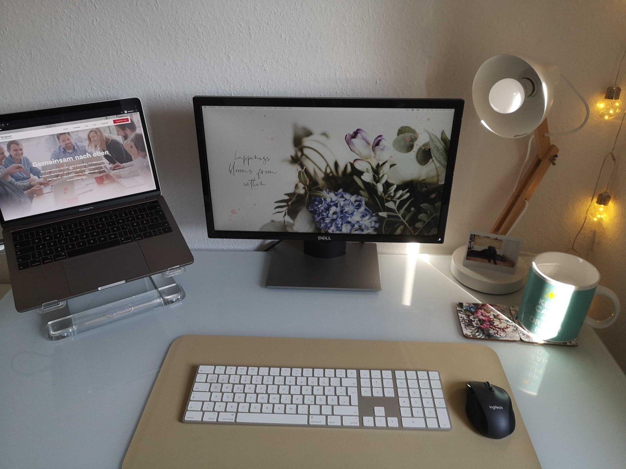 Heller Schreibtisch mit Laptop auf einem Ständer und einem Monitor mit Blumen-Hintergrundbild, dazu Schreibtischlampe, Lichterkette und eine Tasse.