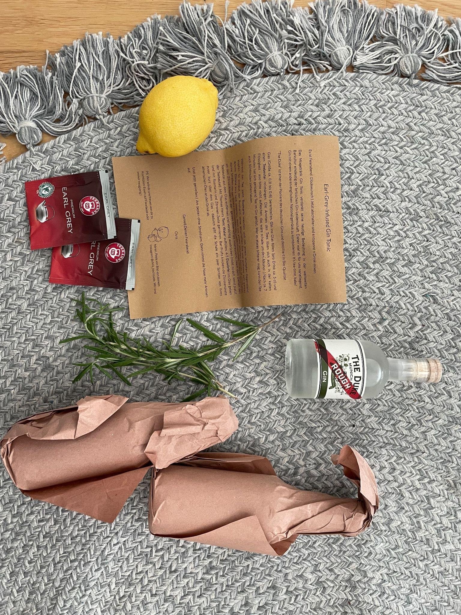 Auf einem grauen Teppich liegen eine Zitrone, zwei Beutel Earl-Grey-Tee, eine Rezeptkarte für einen Earl Grey Infused Gin Tonic, ein Zweig Rosmarin, eine kleine Ginflasche und zwei in Kupferpapier eingeschlagene Päckchen.
