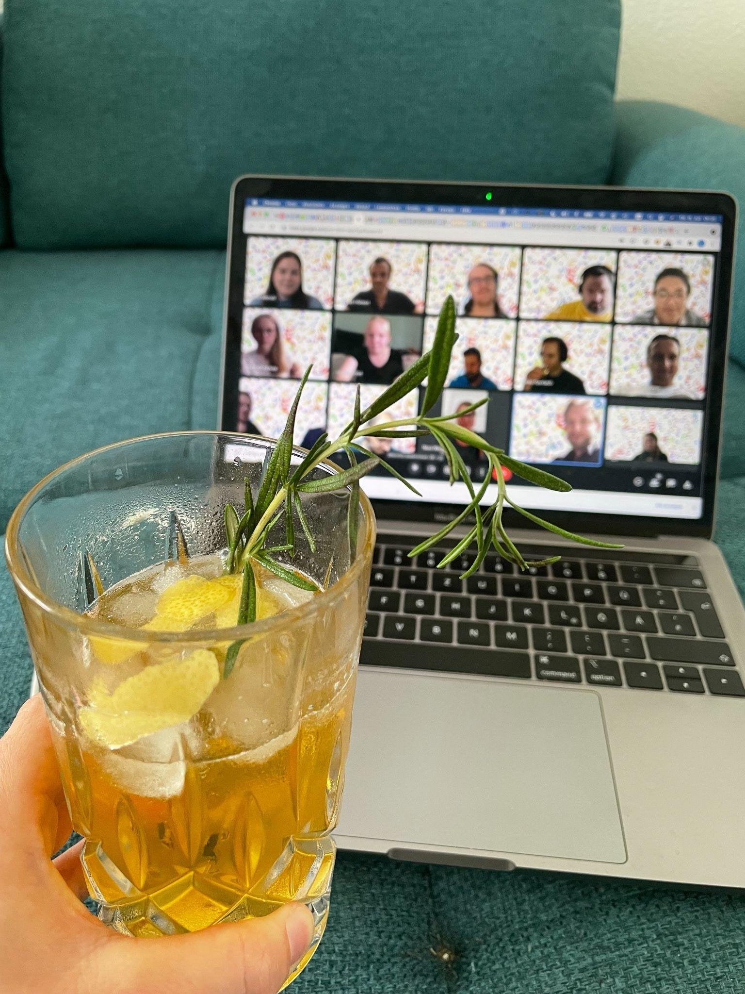 Eine Hand hält ein Glas mit Eis, Zitronenschale und Rosmarin vor einen Laptop, auf dem eine Videokonferenz mit zwölf Teilnehmenden läuft.
