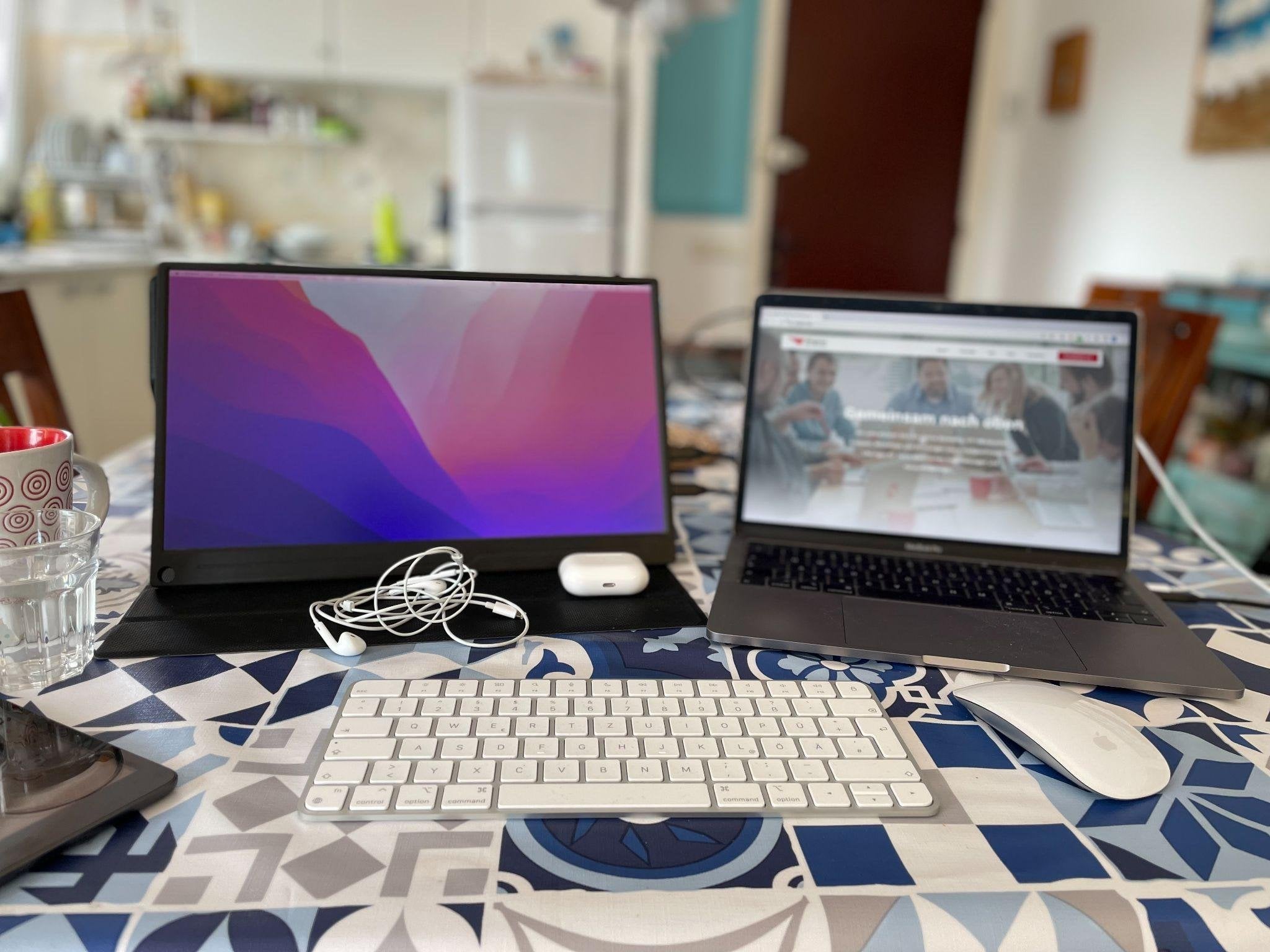 Auf einer gemusterten Tischdecke stehen ein mobiler Zusatzmonitor und ein MacBook mit der Wingmen-Website, davor eine externe Tastatur, eine Maus, Kopfhörer und eine Tasse.
