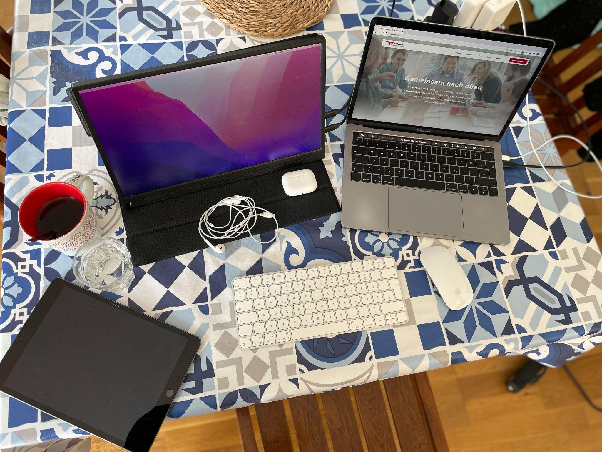 Derselbe Arbeitsplatz von oben fotografiert: Zusatzmonitor, MacBook mit der Wingmen-Website, Tastatur und Maus auf der gemusterten Tischdecke, dazu ein Tablet und ein Glas Wasser.