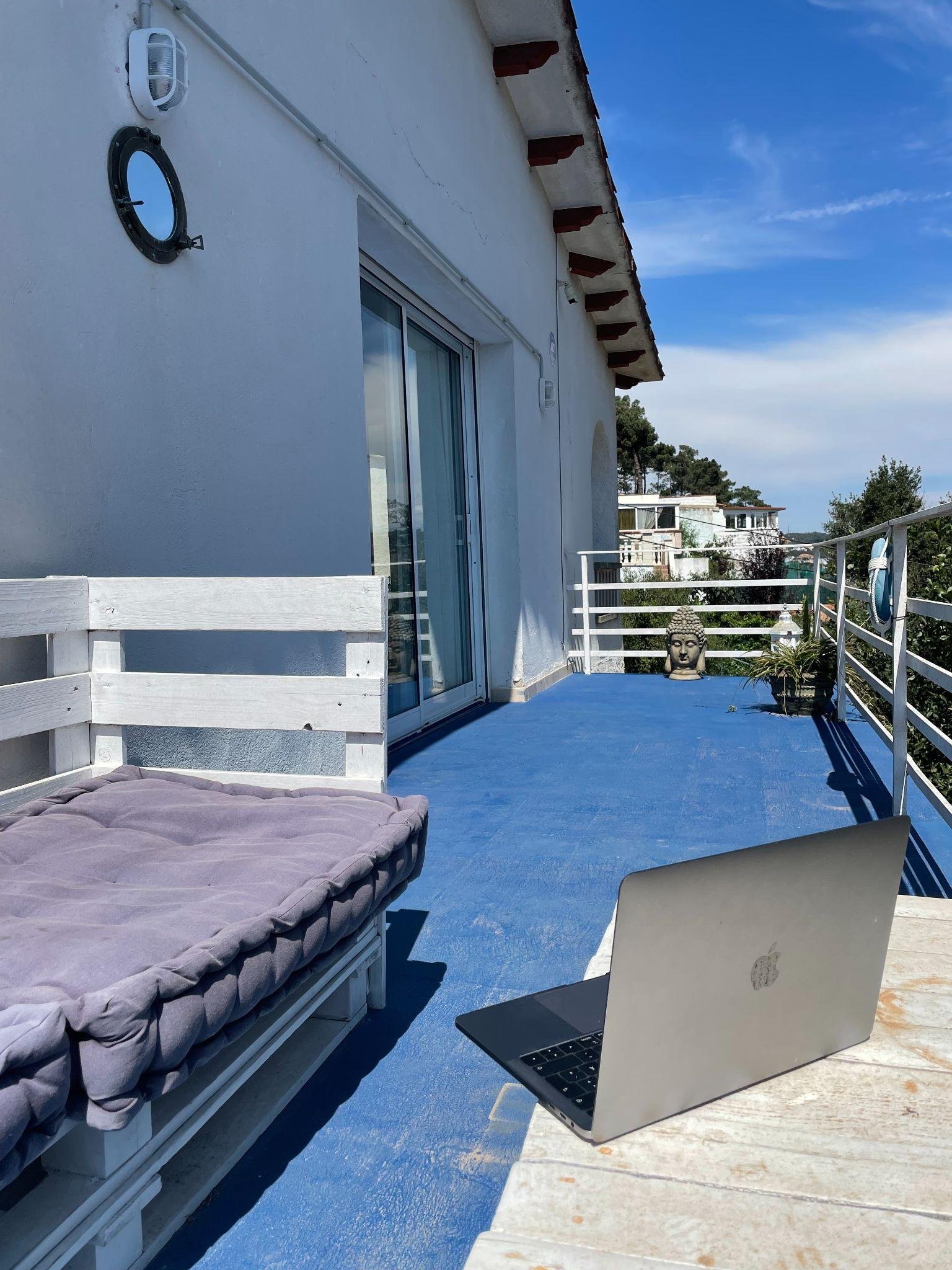 Ein aufgeklappter Laptop steht auf einer blau gestrichenen Terrasse vor einer weißen Hauswand, daneben eine Palettenliege, im Hintergrund Bäume und Häuser.