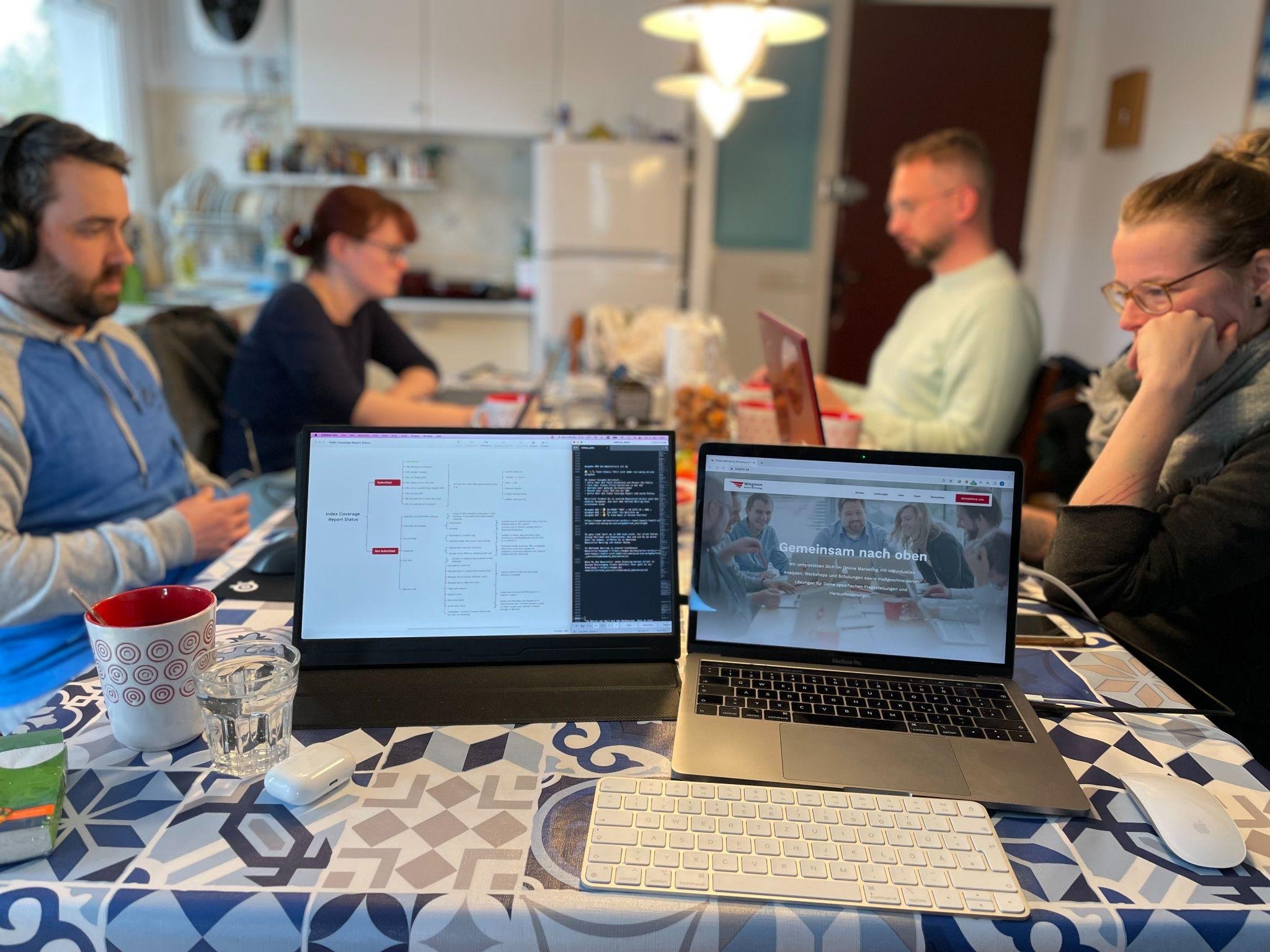 Vier Menschen arbeiten an einem langen Küchentisch, einer davon mit großen Over-Ear-Kopfhörern, im Vordergrund der mobile Zusatzmonitor und das MacBook mit der Wingmen-Website.